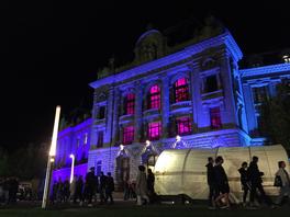 Blau beleuchtetes Hauptgebäude der Universität - aus dem Innern leuchtet es pink.