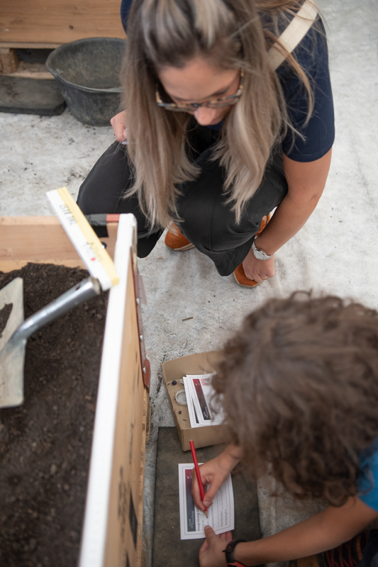 Expertin kniet am Boden, um einem kleinen Archäologen zu erklären, wie der Fundzettel ausgefüllt wird.